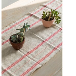 May include: A white linen table runner with red stripes. Two small potted succulents are placed on the runner. The runner has a frayed edge.