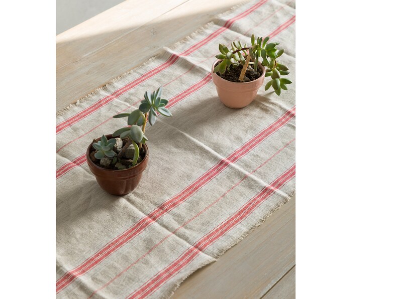 May include: A white linen table runner with red stripes. Two small potted succulents are placed on the runner. The runner has a frayed edge.