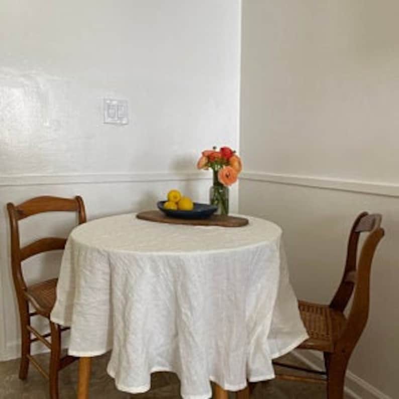May include: A white linen tablecloth with a subtle pattern covers a round wooden table with two wooden chairs. A bowl of lemons sits on a wooden serving tray on the table, and a vase of pink and orange flowers sits on the table.
