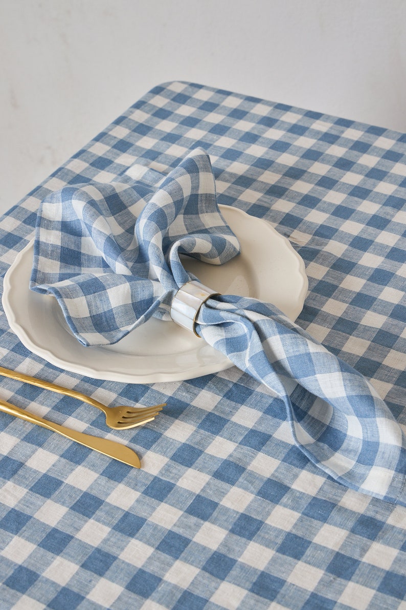May include: A blue and white gingham tablecloth with a white plate and a blue gingham napkin with a gold napkin ring. A gold fork and knife are on the table.