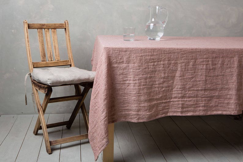 May include: A wooden folding chair with a light grey linen cushion sits next to a table with a pink linen tablecloth. A clear glass jug and a glass of water are on the table.