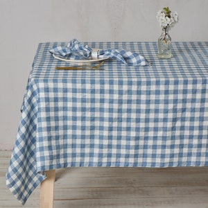 May include: A blue and white gingham linen tablecloth with a white plate and gold silverware on top. The tablecloth is laid out on a wooden table.