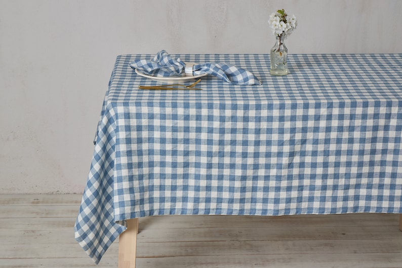 May include: A blue and white gingham linen tablecloth with a white plate and gold silverware on top. The tablecloth is laid out on a wooden table.