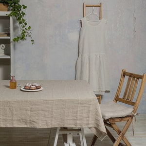 May include: A light brown linen tablecloth draped over a white table with a white wooden easel base. A small plate of pastries sits on the table. A wooden folding chair with a linen cushion sits next to the table. A white linen dress hangs on a wooden ladder.