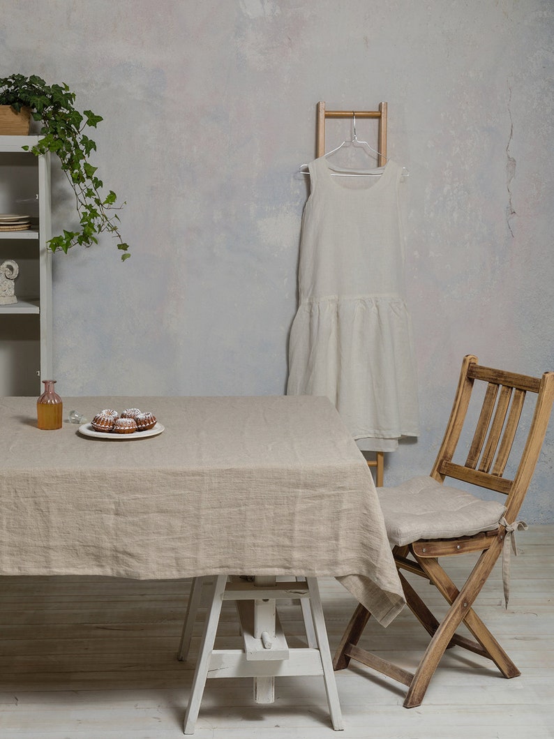 May include: A light brown linen tablecloth draped over a white table with a white wooden easel base. A small plate of pastries sits on the table. A wooden folding chair with a linen cushion sits next to the table. A white linen dress hangs on a wooden ladder.