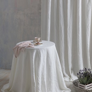 May include: White linen tablecloth draped over a round table with a pink napkin and a teacup and saucer with cookies on a white plate. The table is in front of a white linen curtain and a gray wall.