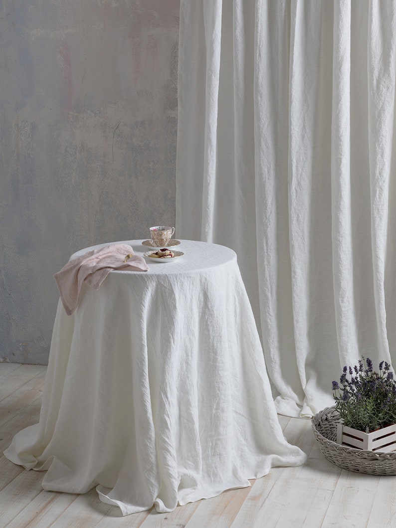 May include: White linen tablecloth draped over a round table with a pink napkin and a teacup and saucer with cookies on a white plate. The table is in front of a white linen curtain and a gray wall.