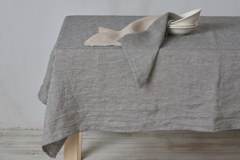 May include: A gray and white striped linen tablecloth with a folded linen napkin on top. The tablecloth is draped over a wooden table.