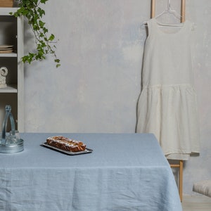 May include: A light blue linen tablecloth with a loaf of bread on a silver tray. The tablecloth is on a table with a metal serving tray and a white dress hanging in the background.