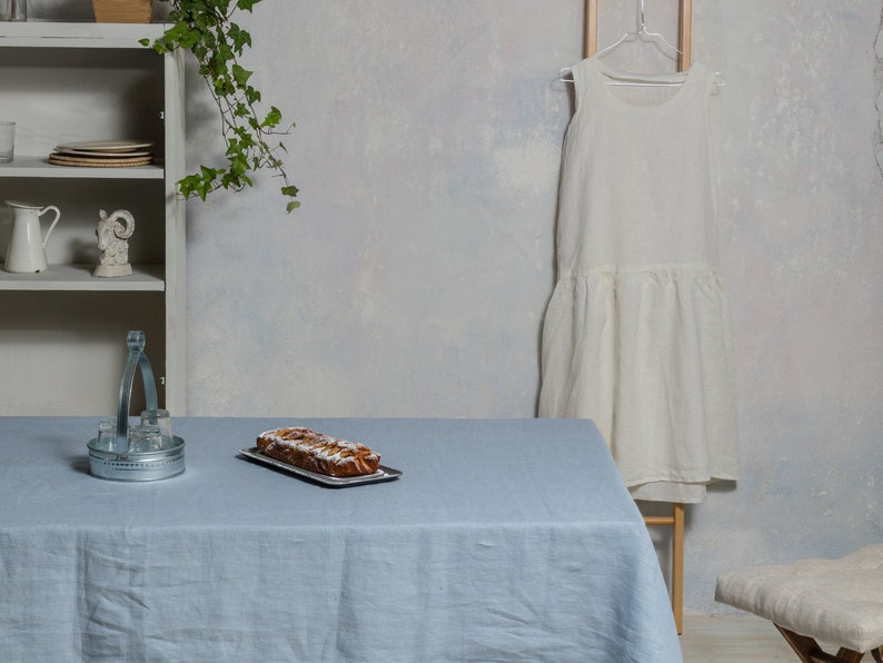May include: A light blue linen tablecloth with a loaf of bread on a silver tray. The tablecloth is on a table with a metal serving tray and a white dress hanging in the background.