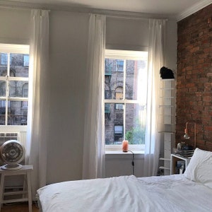 May include: A white bed with white linens and a white headboard is in a room with two windows with white curtains. The windows look out onto a brick building. There is a white shelf unit to the right of the bed.