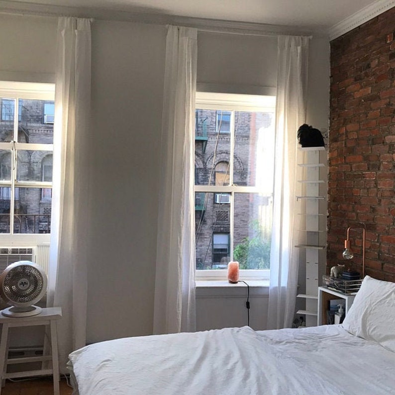 May include: A white bed with white linens and a white headboard is in a room with two windows with white curtains. The windows look out onto a brick building. There is a white shelf unit to the right of the bed.