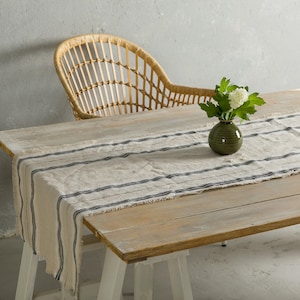 May include: A wooden table with a white and black striped linen table runner. A green vase with white flowers sits on the table runner. A wicker chair is in the background.