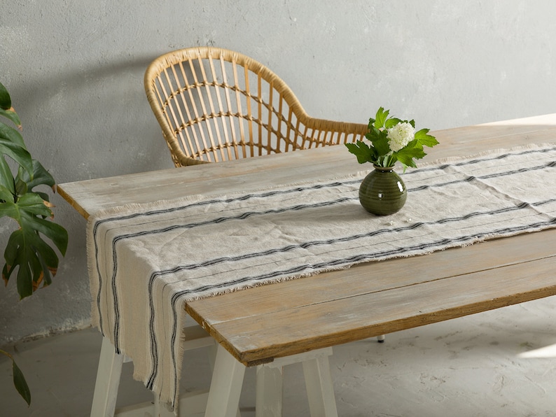 May include: A wooden table with a white and black striped linen table runner. A green vase with white flowers sits on the table runner. A wicker chair is in the background.