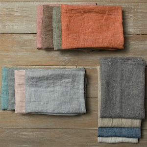 May include: A set of linen kitchen towels in various colors, including teal, pink, gray, orange, and brown. The towels are folded and stacked on a wooden surface.