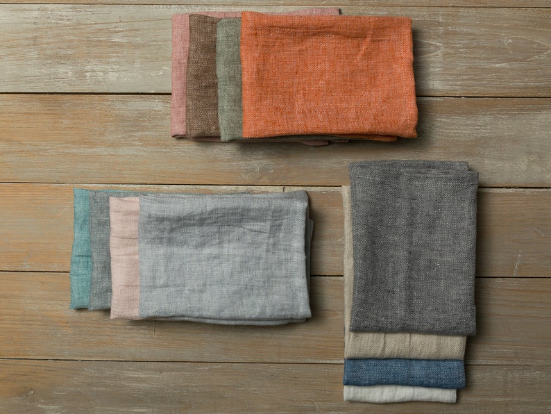 May include: A set of linen kitchen towels in various colors, including teal, pink, gray, orange, and brown. The towels are folded and stacked on a wooden surface.