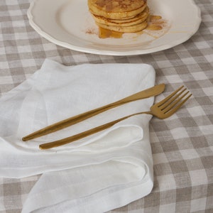 May include: A white linen napkin folded on a brown and white checkered tablecloth with two gold utensils on top.