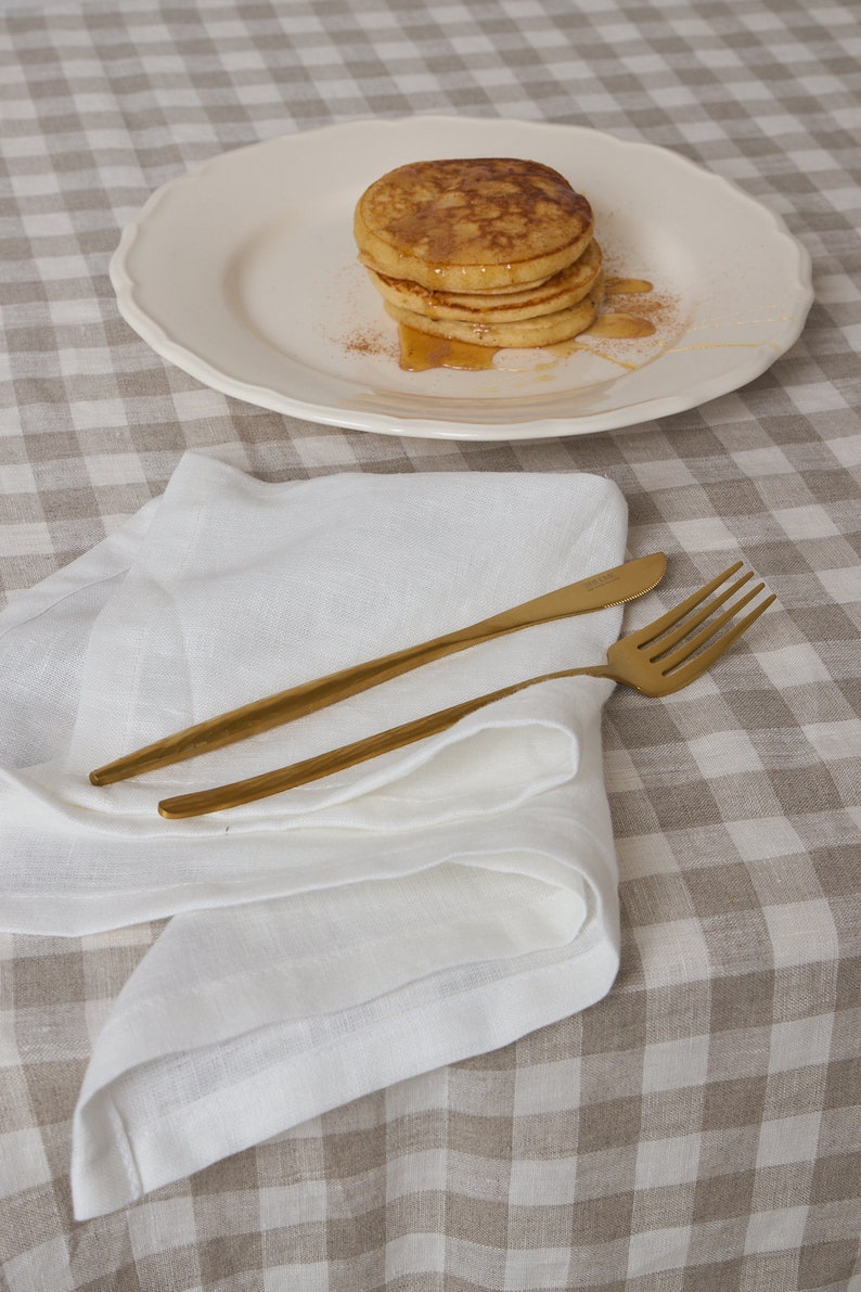 May include: A white linen napkin folded on a brown and white checkered tablecloth with two gold utensils on top.