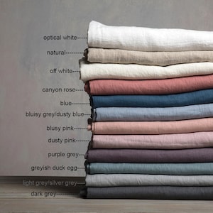 Op de afbeelding: Een stapel van 12 linnen stofstalen in verschillende kleuren, waaronder optisch wit, naturel, off white, canyon rose, blauw, stoffig blauw, stoffig roze, paars grijs, grijsachtig eendenei, licht grijs en donker grijs.