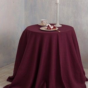 Puede incluir: Un mantel redondo de lino color burdeos drapeado sobre una mesa con un candelero blanco, un plato peque&ntilde;o con un postre y una taza de t&eacute; y un platillo.