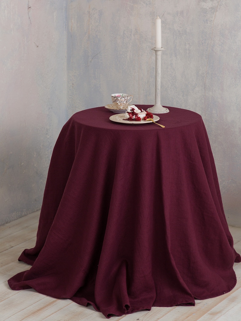 Puede incluir: Un mantel redondo de lino color burdeos drapeado sobre una mesa con un candelero blanco, un plato peque&ntilde;o con un postre y una taza de t&eacute; y un platillo.