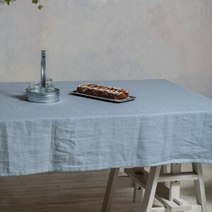 May include: A light blue linen tablecloth covers a wooden table with a white wooden easel underneath. A loaf of bread with a brown crust and a white topping is on a silver tray on the table. A silver metal container with a handle is on the table.