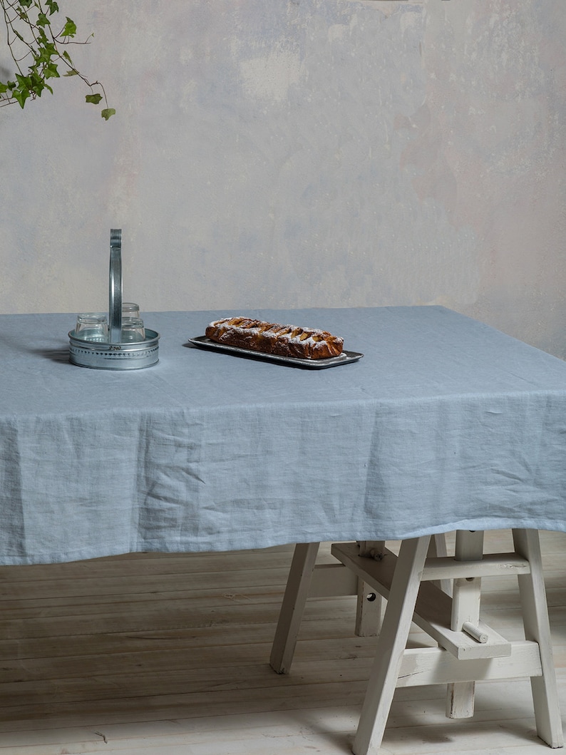 May include: A light blue linen tablecloth covers a wooden table with a white wooden easel underneath. A loaf of bread with a brown crust and a white topping is on a silver tray on the table. A silver metal container with a handle is on the table.