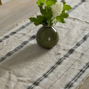 May include: A green ceramic vase with white flowers sits on a white and black striped table runner.