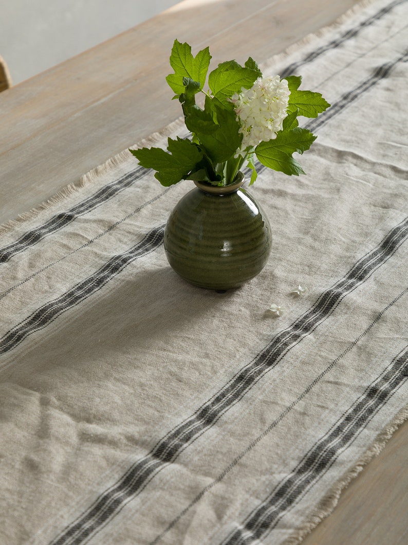 May include: A green ceramic vase with white flowers sits on a white and black striped table runner.