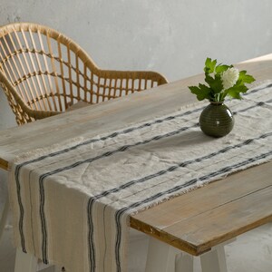 May include: A white and black striped linen table runner with frayed edges on a wooden table with a white vase of white flowers.