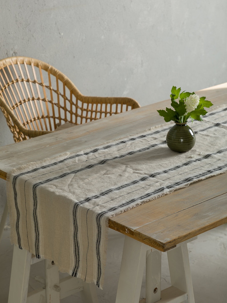 May include: A white and black striped linen table runner with frayed edges on a wooden table with a white vase of white flowers.