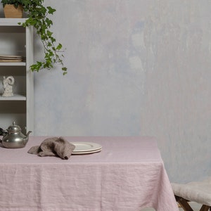 May include: A pink linen tablecloth with a silver teapot, a brown linen napkin, and a white plate on a wooden table. A wooden chair with a white cushion is in the background.