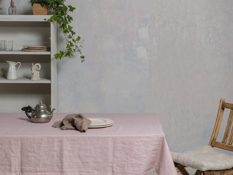 May include: A pink linen tablecloth with a silver teapot, a brown linen napkin, and a white plate on a wooden table. A wooden chair with a white cushion is in the background.