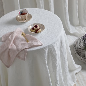 May include: A white linen tablecloth with a pink linen napkin, a gold spoon, a teacup and saucer with floral pattern, and a small plate with four cookies on a white table.