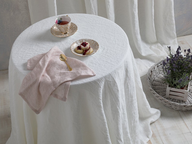 May include: A white linen tablecloth with a pink linen napkin, a gold spoon, a teacup and saucer with floral pattern, and a small plate with four cookies on a white table.