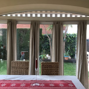 May include: A white table with a red and white snowflake patterned table runner. Two wicker chairs are placed on either side of the table. A single red candle sits in a clear glass holder on the table. The table is set in front of a window with sheer curtains and a view of a lush green garden.
