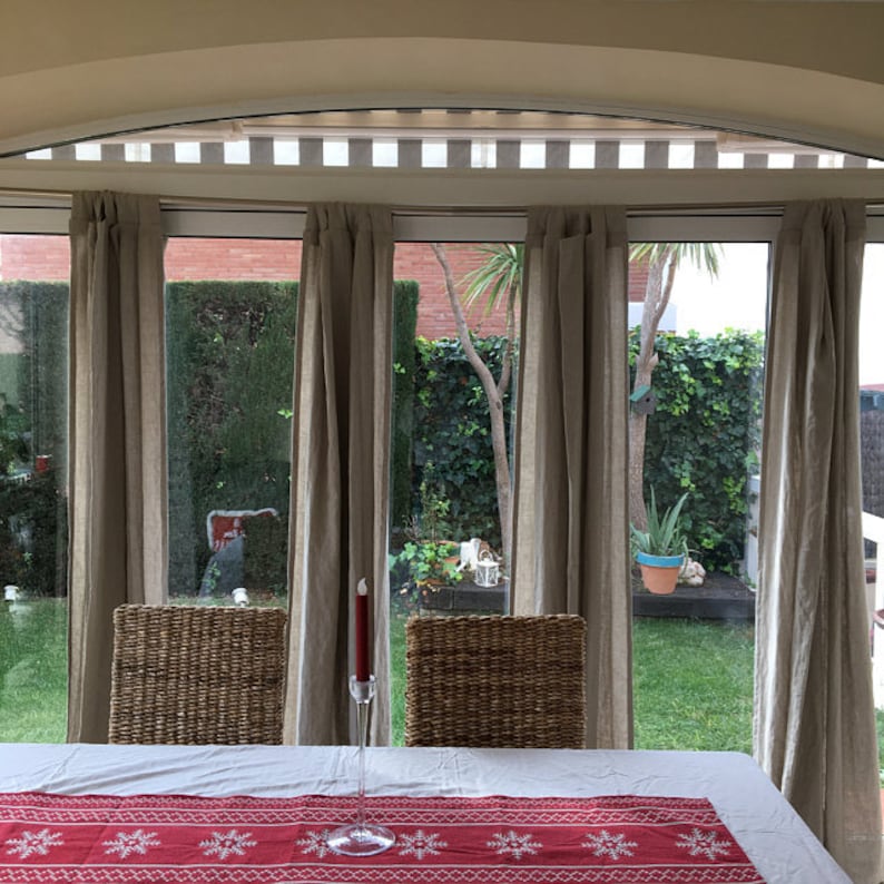 May include: A white table with a red and white snowflake patterned table runner. Two wicker chairs are placed on either side of the table. A single red candle sits in a clear glass holder on the table. The table is set in front of a window with sheer curtains and a view of a lush green garden.