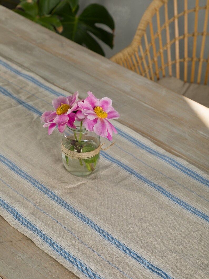 May include: A light brown table runner with blue stripes, featuring a glass jar with pink flowers on top.