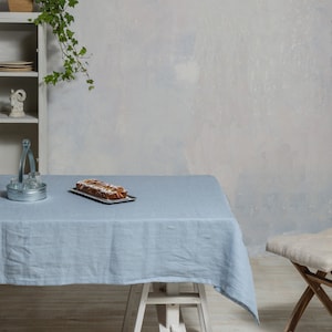 May include: A light blue linen tablecloth draped over a white wooden table with a loaf of bread on a plate. The table is set against a white wall with a potted plant in the background.