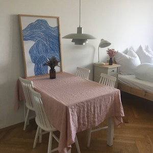 May include: A pink linen tablecloth covers a white dining table with four white chairs. A framed blue and white abstract print hangs on the wall behind the table. A grey pendant light hangs above the table.