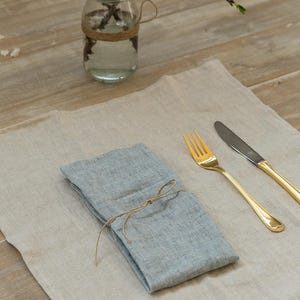 May include: A light blue linen napkin tied with twine, placed on a beige linen placemat with a gold fork and knife.