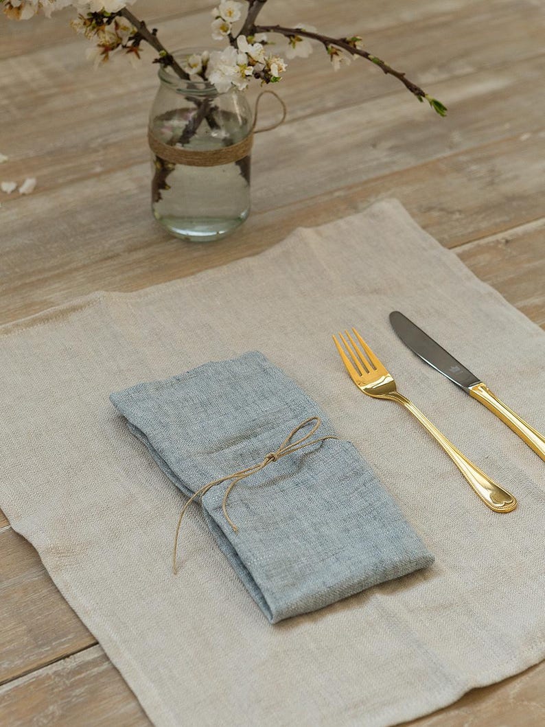 May include: A light blue linen napkin tied with twine, placed on a beige linen placemat with a gold fork and knife.