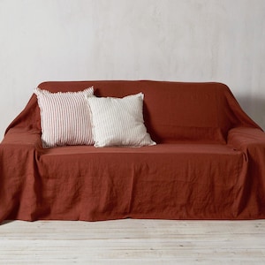 Op de afbeelding: Een linnen sofa-overtrek in roestkleur met twee decoratieve kussens, een met witte strepen en een met rode strepen.
