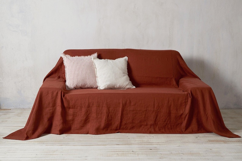 Op de afbeelding: Een linnen sofa-overtrek in roestkleur met twee decoratieve kussens, een met witte strepen en een met rode strepen.