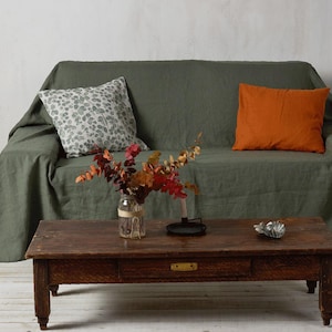 Op de afbeelding: Een groene linnen bank met twee kussens, een met een wit en groen bloemenpatroon en de andere effen oranje. De bank heeft een houten salontafel ervoor met een vaas met gedroogde bloemen en een decoratief object.
