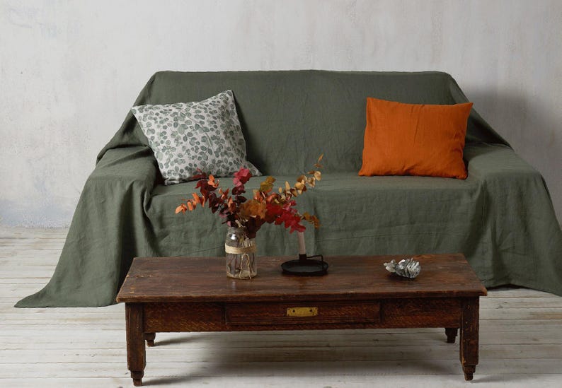 Op de afbeelding: Een groene linnen bank met twee kussens, een met een wit en groen bloemenpatroon en de andere effen oranje. De bank heeft een houten salontafel ervoor met een vaas met gedroogde bloemen en een decoratief object.