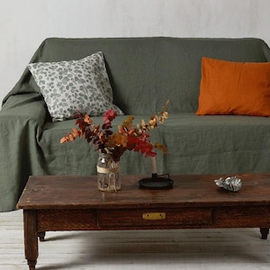 Op de afbeelding: Een groene linnen bank met twee kussens, een met een wit en grijs bladmotief en de andere effen oranje. Een houten salontafel staat voor de bank met een vaas met gedroogde bloemen.