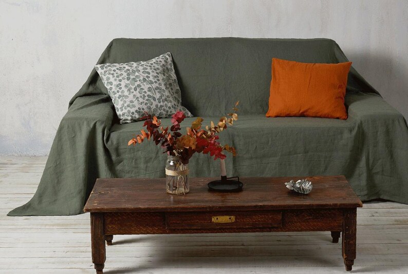 Op de afbeelding: Een groene linnen bank met twee kussens, een met een wit en grijs bladmotief en de andere effen oranje. Een houten salontafel staat voor de bank met een vaas met gedroogde bloemen.