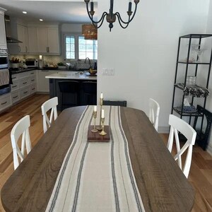 May include: A wooden dining table with a white and black striped runner. The table is set with four white chairs and three gold candle holders with white candles.