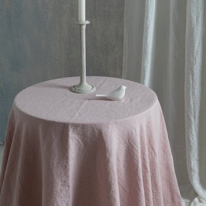 Puede incluir: Un mantel de lino rosa drapeado sobre una mesa redonda con un p&aacute;jaro de cer&aacute;mica blanca y una vela blanca en un candelero de metal gris.